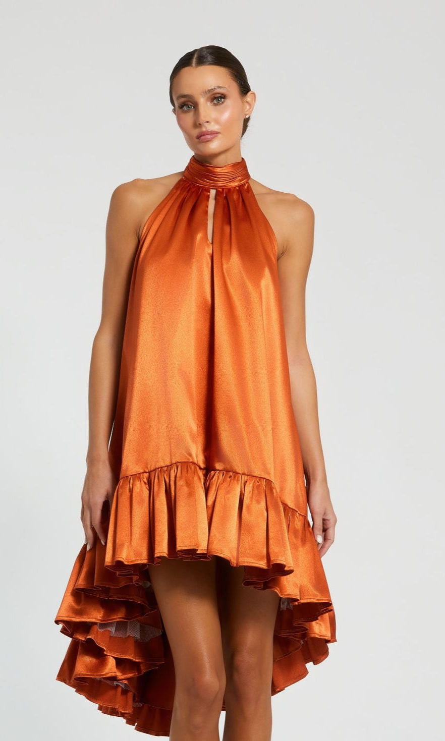 A woman models the Mac Duggal 20711 Satin Wedding Guest Dress—sleeveless, high-neck, and featuring a ruffled high-low hem—against a plain light background.